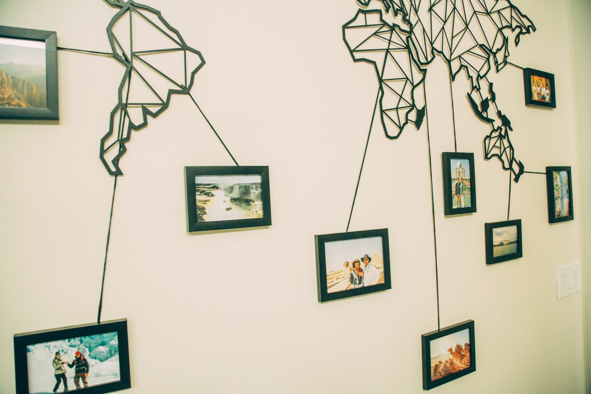 A closeup look at the travel wall, zoomed in on picture frames connected to a metal world map mounted on a beige wall. The photos are linked to their locations around the world using a simple strand of string.