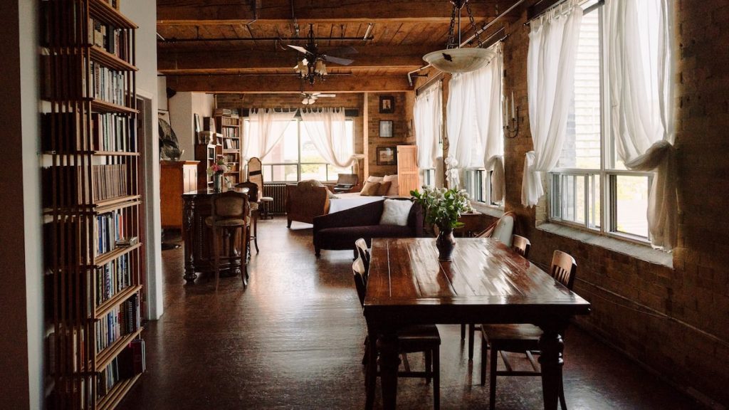 A rustic style studio loft features exposed wood beam ceilings and dark natural hardwood floors, a dining room, and living room area. The walls are lined with bookshelves and crisp white linens drape over the windows.