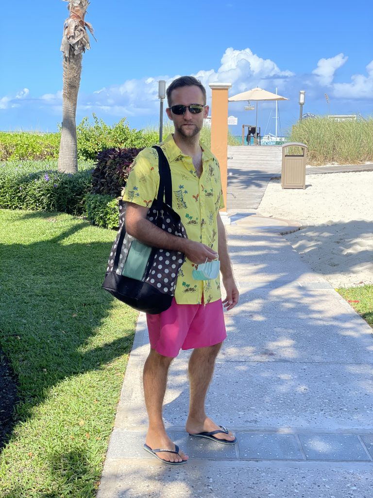 A man in Hawaiian shirt and a pink shorts carrying a polka dot beach bag.