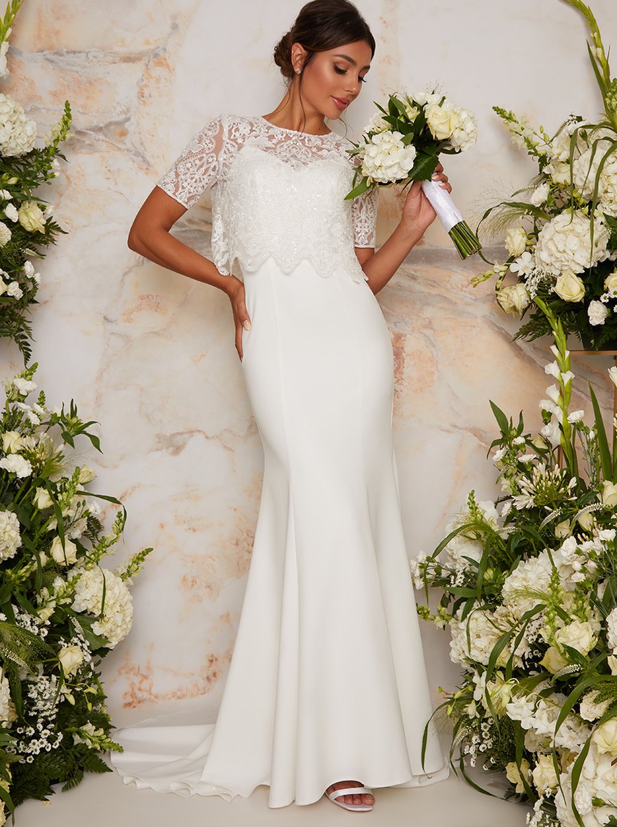 A model wears a long white maxi dress with a lace short-sleeved bodice. The dress has a sweetheart neckline and short train. The model is surrounded by white flower arrangements and holds a white bridal bouquet.
