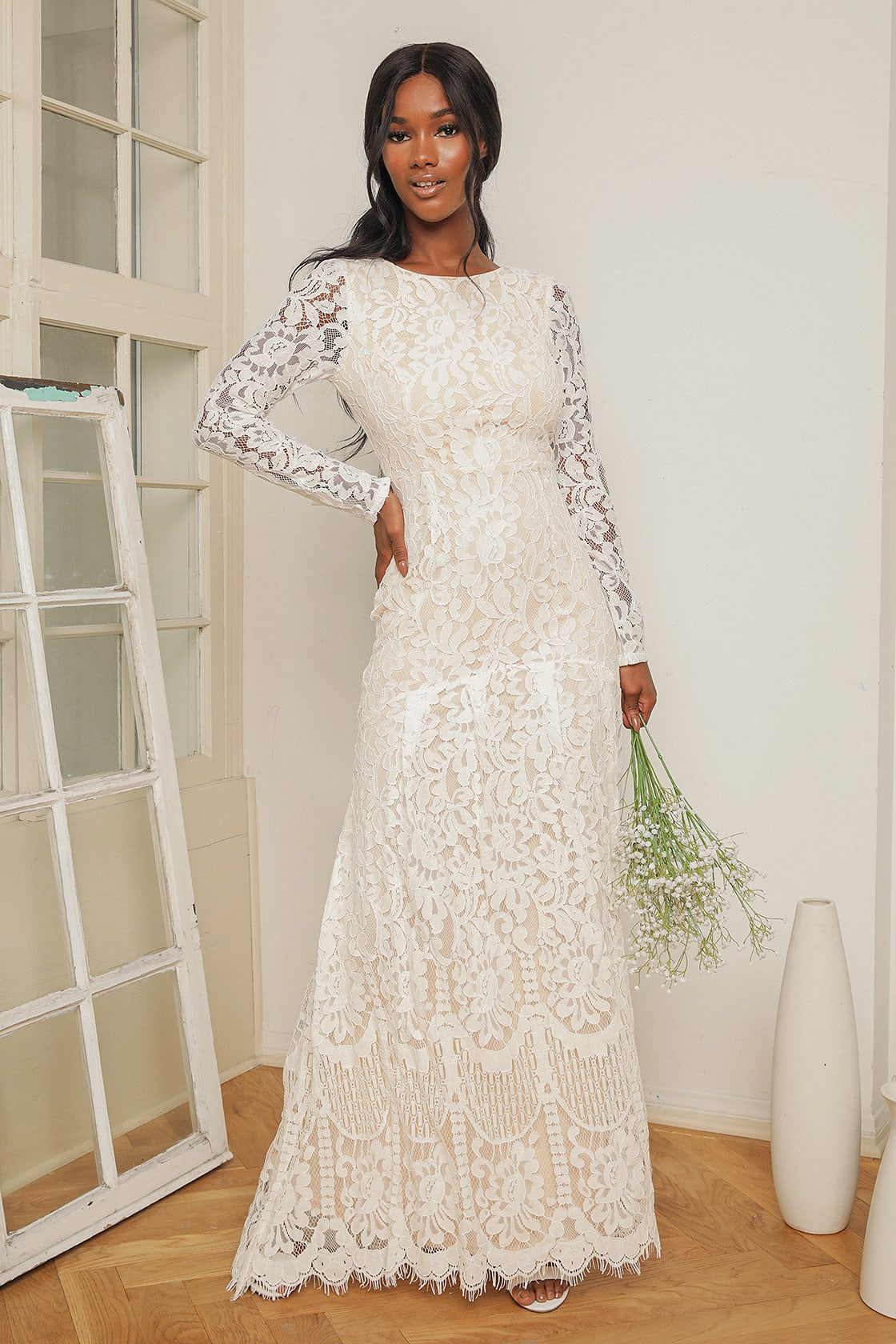 A model wears a long-sleeved lace wedding dress. The white dress is mermaid style with an all-over lace fabric that extends up the bodice with a high neckline and down the sleeves.