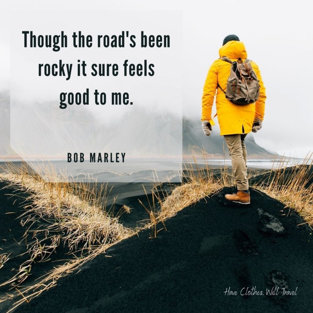 A person in a yellow coat stands on a rock, overlooking a lake and foggy mountain range. Text across the image reads, "Though the road's been rocky, it sure feels good to me. - Bob Marley"