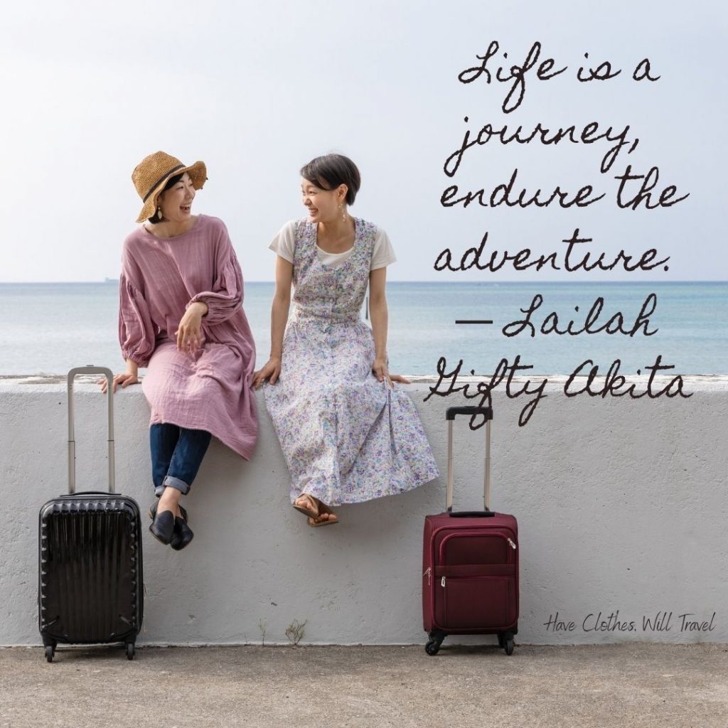 Two women sit on a white stone wall, looking at each other and laughing; their suitcases sit beside them. Text across the image reads, 