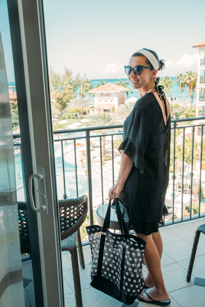 Lindsey wearing black sunglasses and a black swimsuit cover up that ties in the back and her hair in a ponytail with a white headband. She is holding a polka dot beach bag and standing on a balcony at the Beaches Turks and Caicos resort