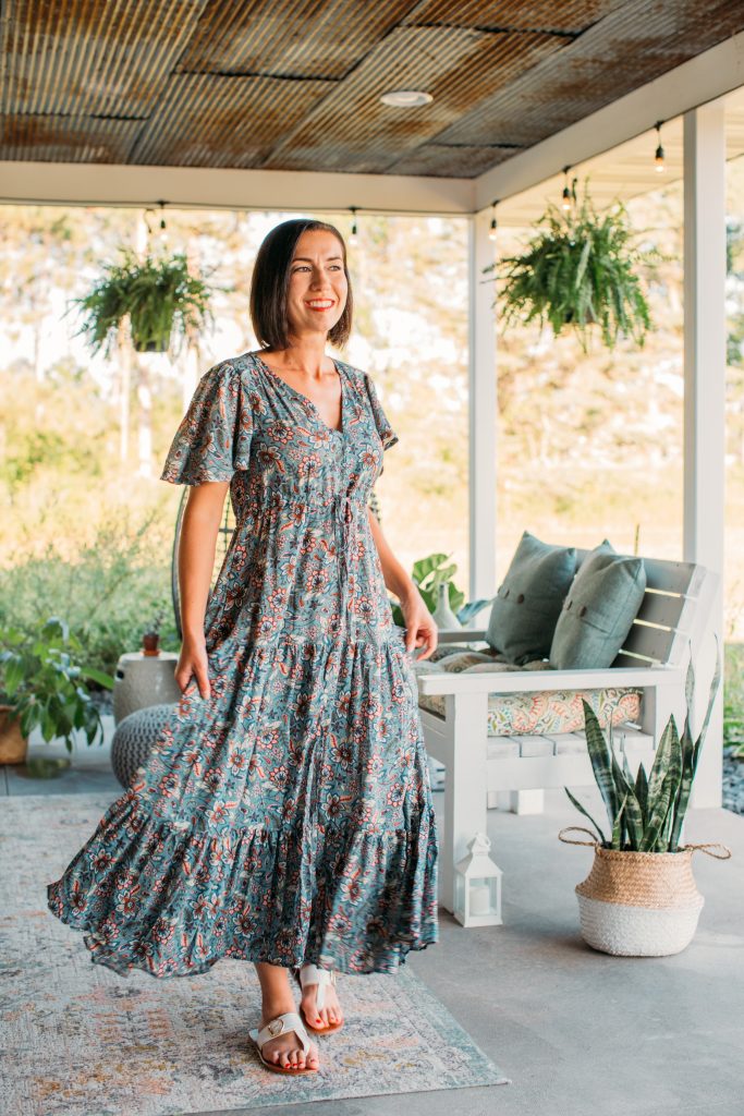 Lindsey wearing a Salty Marla Maxi Dress - Teal standing on a covered porch with hanging ferns and patio furniture