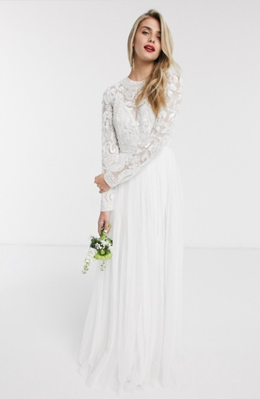 A model wears a long, floor length white modest wedding dress. The bodice is beaded with shimmering silver beading that extends all the way down the long sleeves. The tulle skirt is long and flowy, just touching the floor.