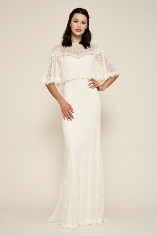A model wears a long floor length white wedding dress with a sleek lace skirt and cropped lace overlay bodice. The flutter sleeves give the dress a vintage vibe and the sweetheart neckline is simple and modest.