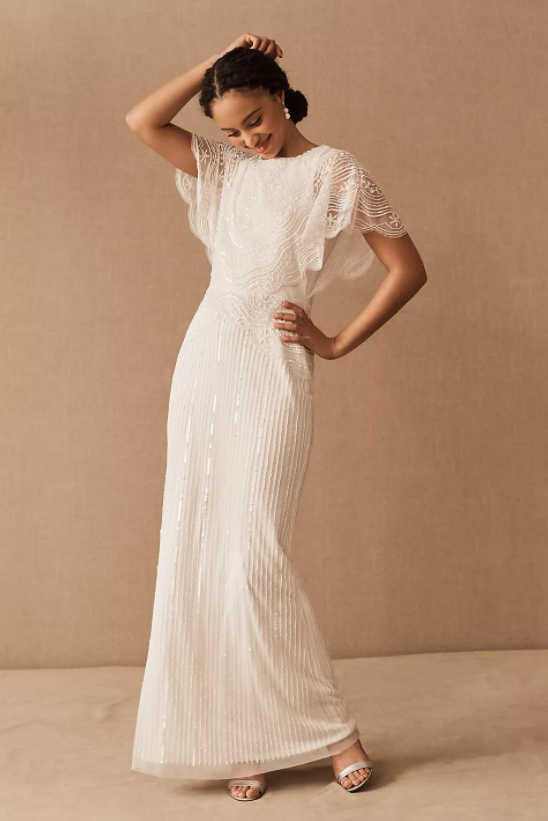 A model poses in a long, floor length white wedding dress. The body of the dress has long rows of shimmering beads and sequins, and the sleeves are short and flowy.