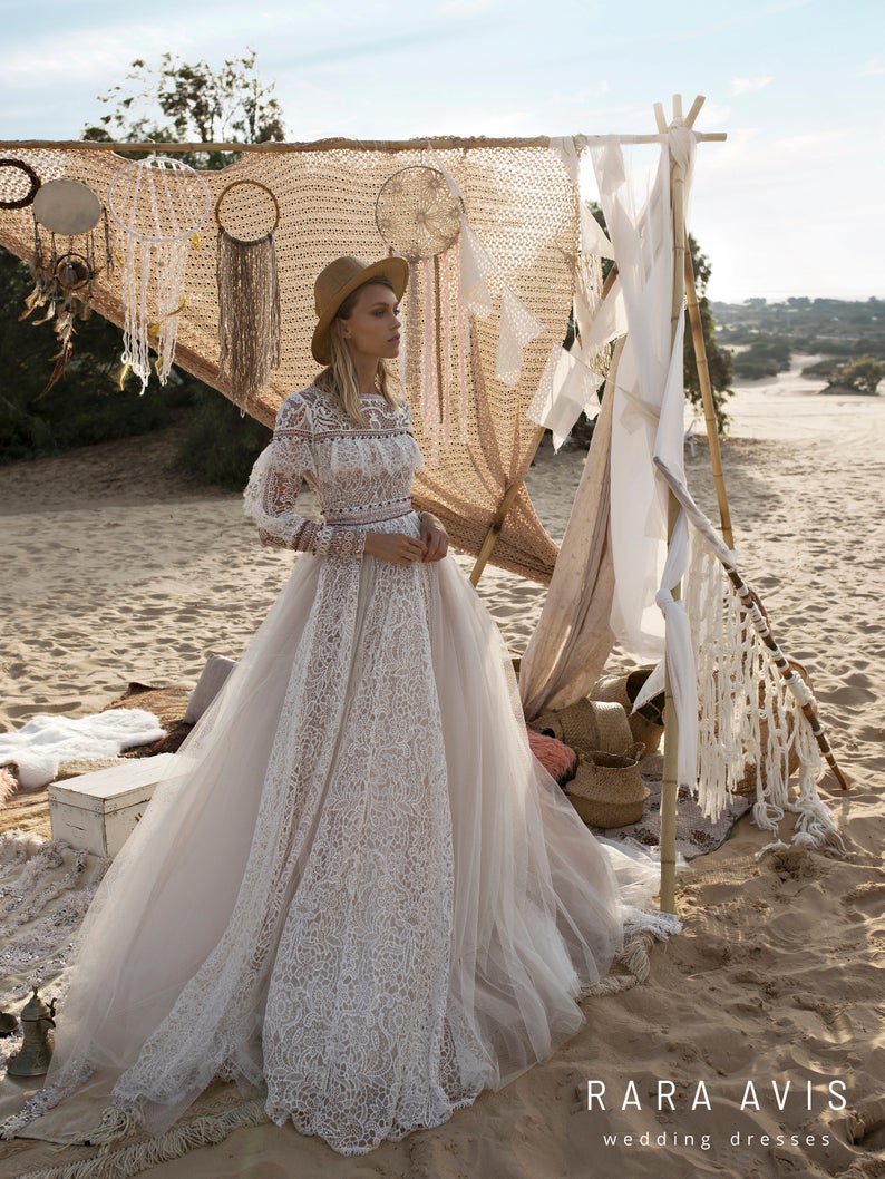 A model is wearing a modest bohemian style wedding dress. The dress has a long, full length tulle skirt with a stunning lace pattern. The bodice is long sleeve lace with a high neckline. The background is a sandy dessert scene with a boho backdrop decorated with linens and dreamcatchers.