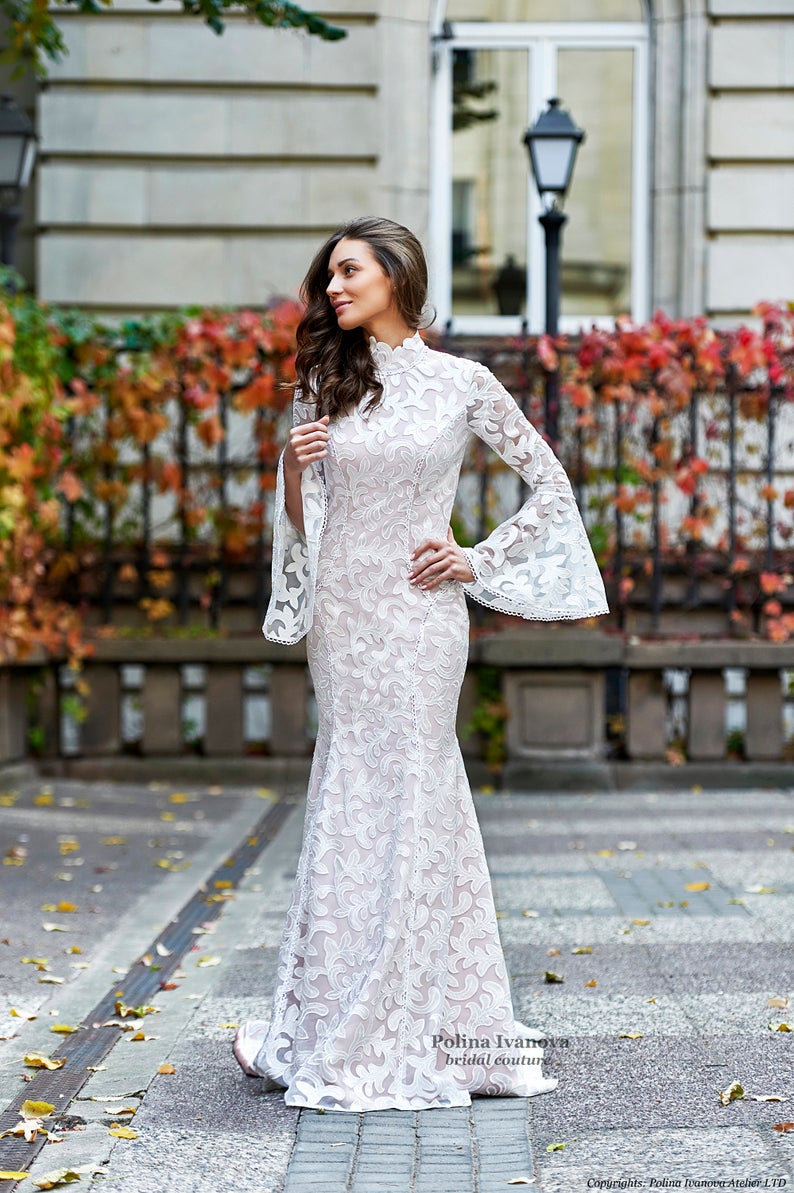 A model wears a long, modest wedding dress by Polina Ivanova. The stress has an all-over embroidered flower pattern, long lace bell sleeves, and a high mock neck neckline.