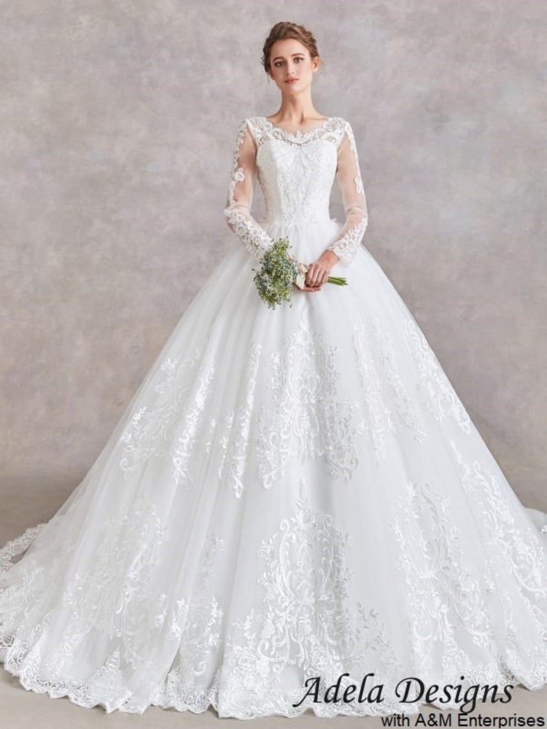 A model wears a floor length white ballgown wedding dress. The large, full skirt is detailed with a shimmering fabric. The bodice of the dress has long lace sleeves, a sweetheart neckline, and lace collar.
