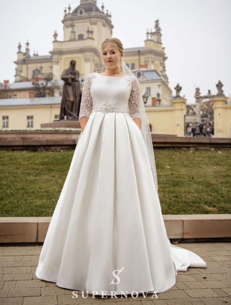 A model wears a classic style white wedding dress. The skirt is a smooth silk fabric that extends to the ground with a long train. The bodice has lace detailing at the waist, and modest lace three-quarter length sleeves. The model is also wearing a long veil.