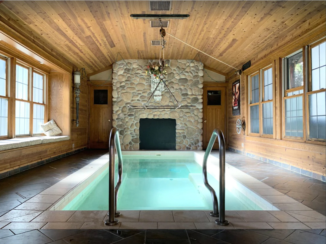 An indoor swimming pool inside a cabin, with two walls of window and a stone fireplace.