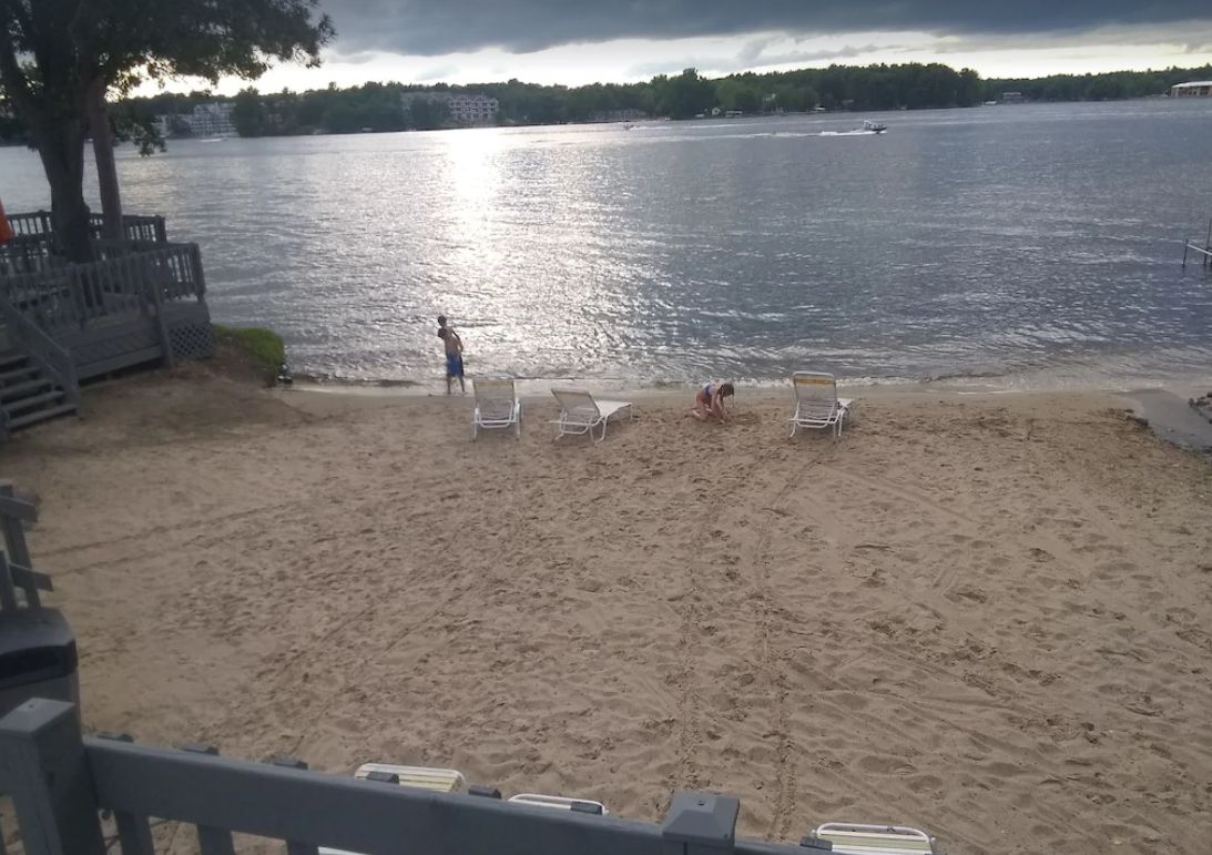 A picture of the lakefront taken from the balcony of a Wisconsin Dells cabin rental. A private sandy beach leads to the calm waters. Two children play in the sand near the waterline.