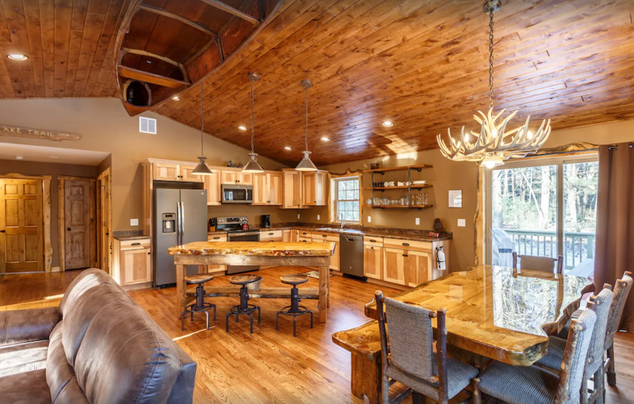 An interior image of a large cabin rental in the Wisconsin Dells shows an open concept living room, kitchen area, and  dining room. The decor and furniture is all natural wood and neutral colors. The vaulted ceiling has a canoe mounted upside down, and a deer antler chandelier hanging over the dining table.