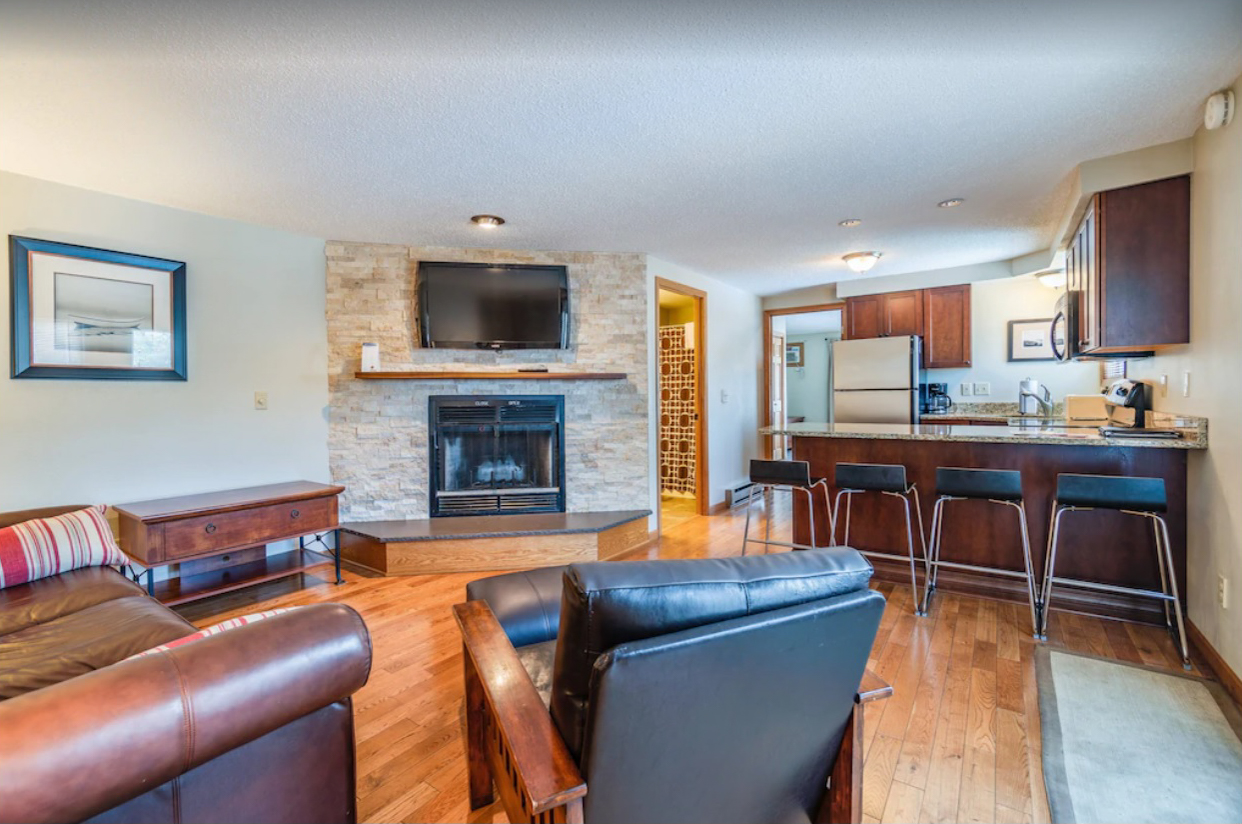 An interior image of the living room and dining room areas in a Lake Delton condo rental. A leather couch and chair face a fireplace and flat screen TV. A breakfast bar is attached to a small kitchen, all with natural light from large windows.