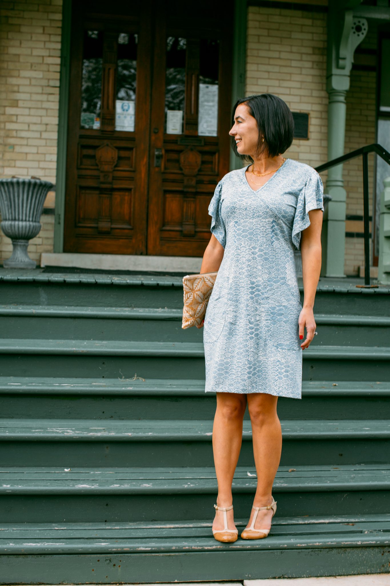 A woman poses standing on green painted stairs of a house. She's wearing a stylish aqua blue patterned wrap dress, and looking over her shoulder away from the camera.