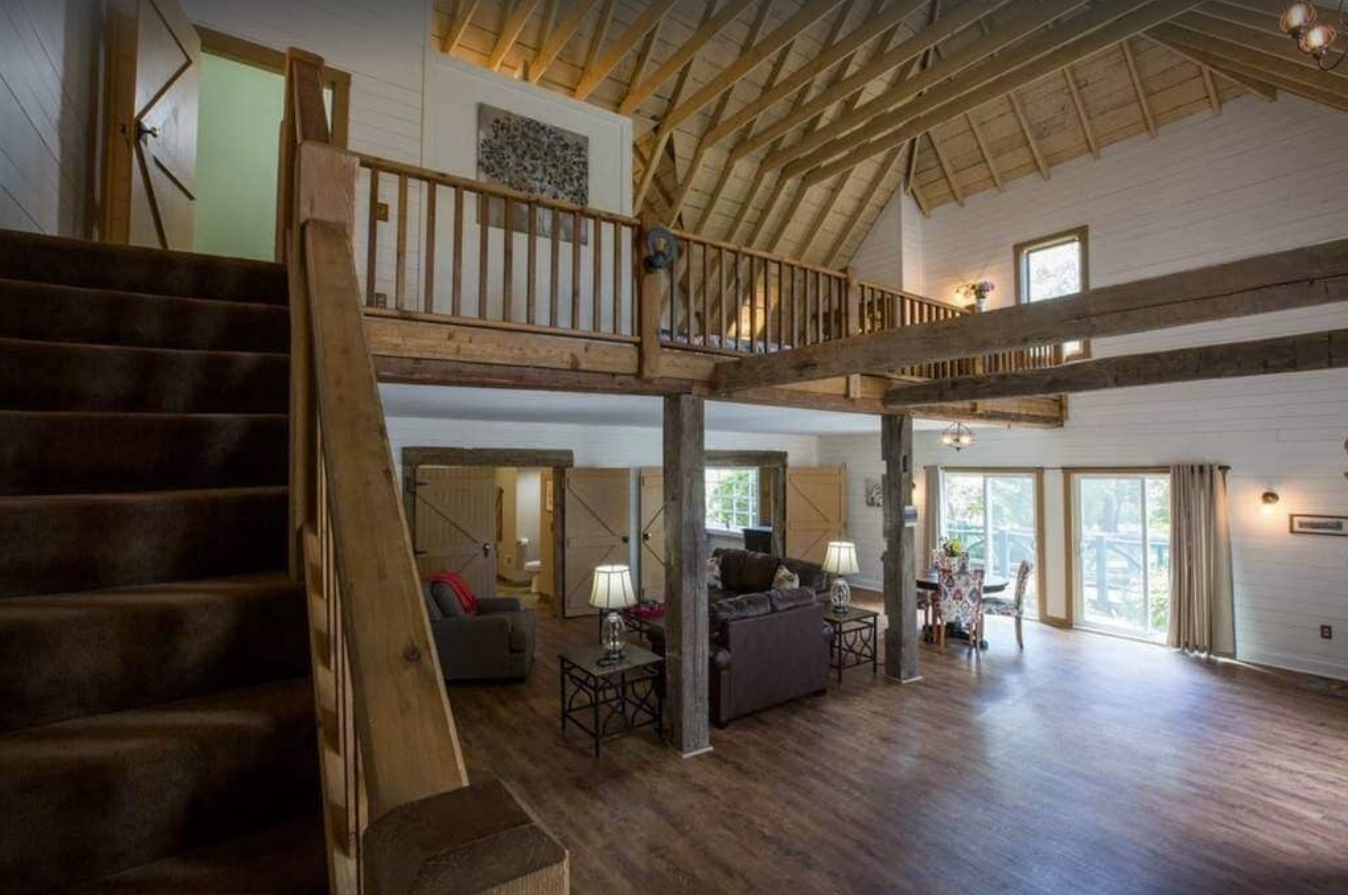 An interior image of a renovated coach house vacation rental in Door County. The cabin-style home is two stories and features a loft style upper bedroom and balcony railing. On the first level is an open concept living room with large windows, hardwood floors, and barn-style doors.
