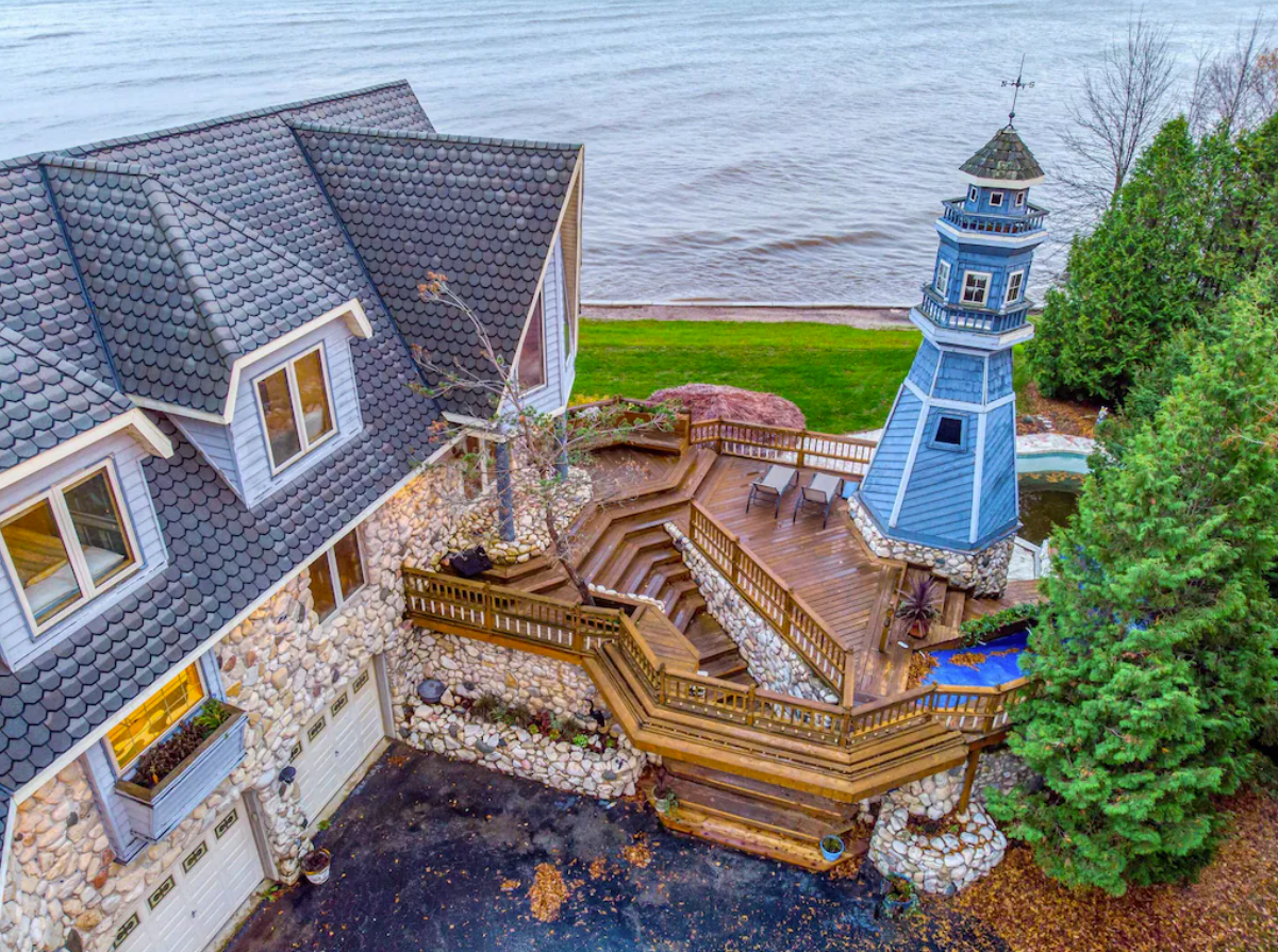 An aerial image of a large lakefront house in Door County, WI. The backyard has an expansive wood deck featuring a pool and blue light house. The exterior of the home is all stone with a unique shingled roof.