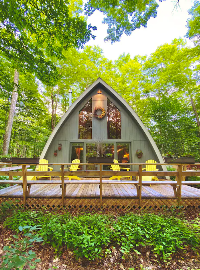 An exterior image of the back porch and yard of an a-frame cabin in the woods of Door County. The back of the small cabin is filled with windows and a large wood deck with yellow Adirondack chairs.