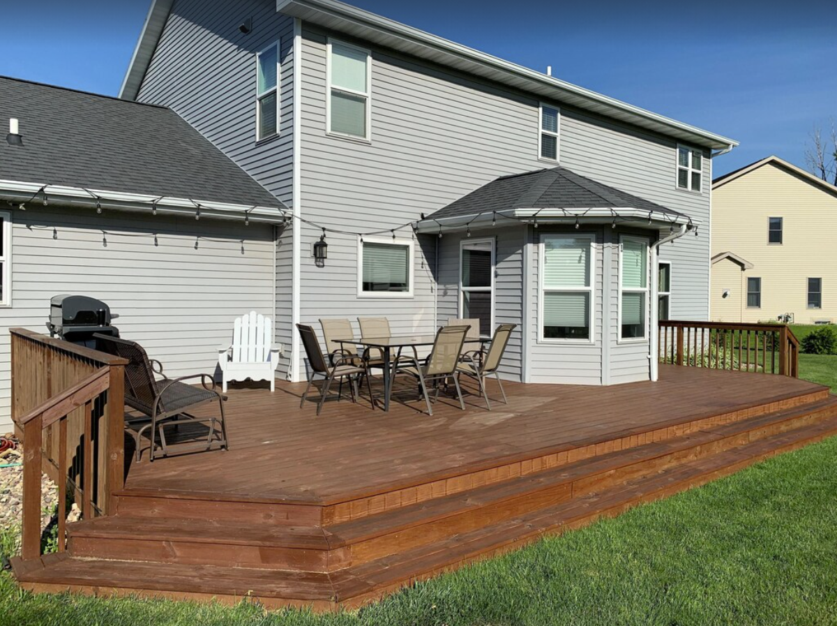 An exterior shot of the back porch of a two-story home with a deck. The home has gray siding and a dark asphalt shingle roof. The wooden deck is decorated with patio furniture and string lights hanging along the gutters of the home.