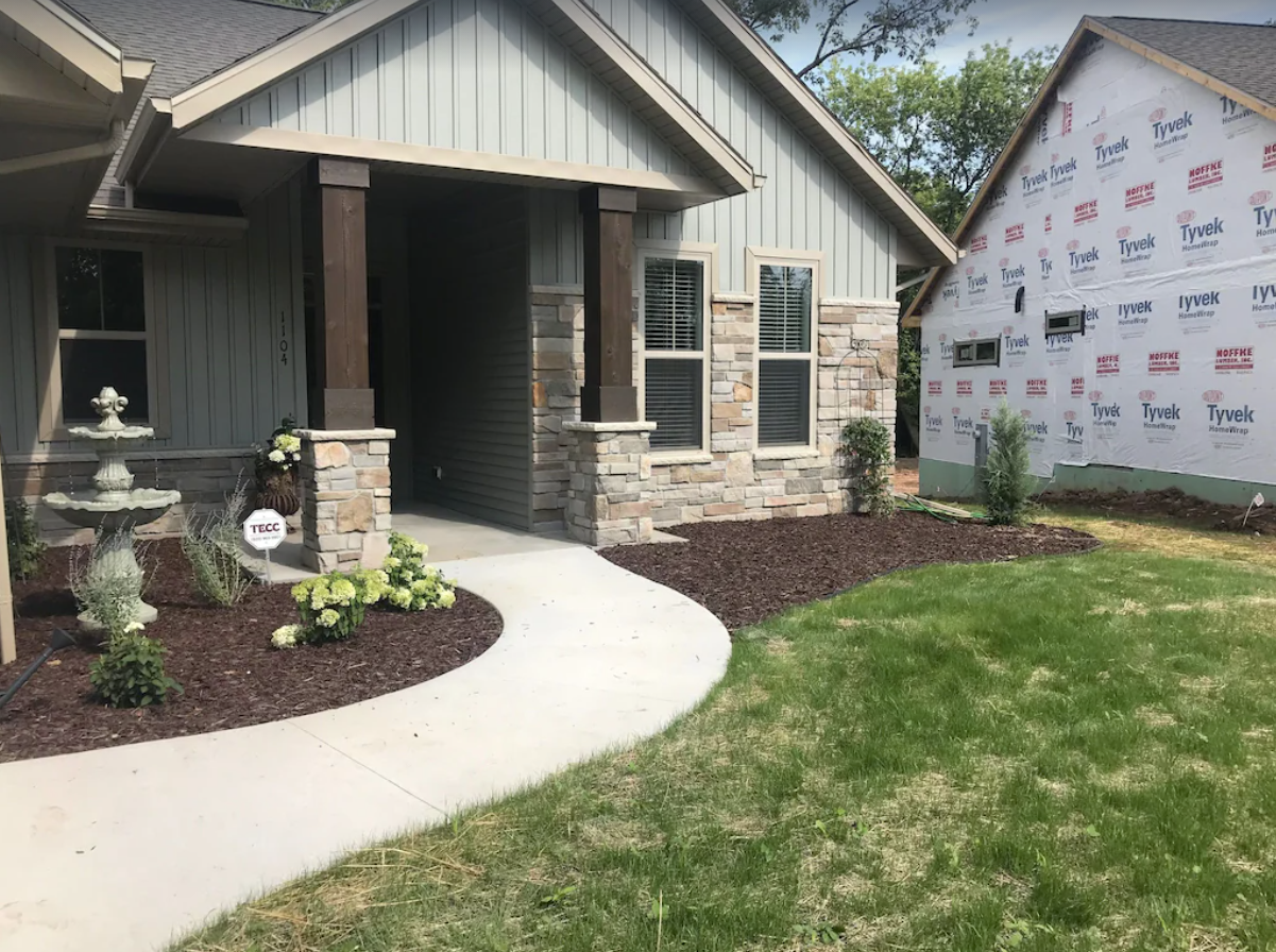 An exterior shot of the front entrance of a home with gray siding and multi-colored natural stone walls. Flower beds with brown mulch line the front of the home, with flowers, plants, and a birdbath.