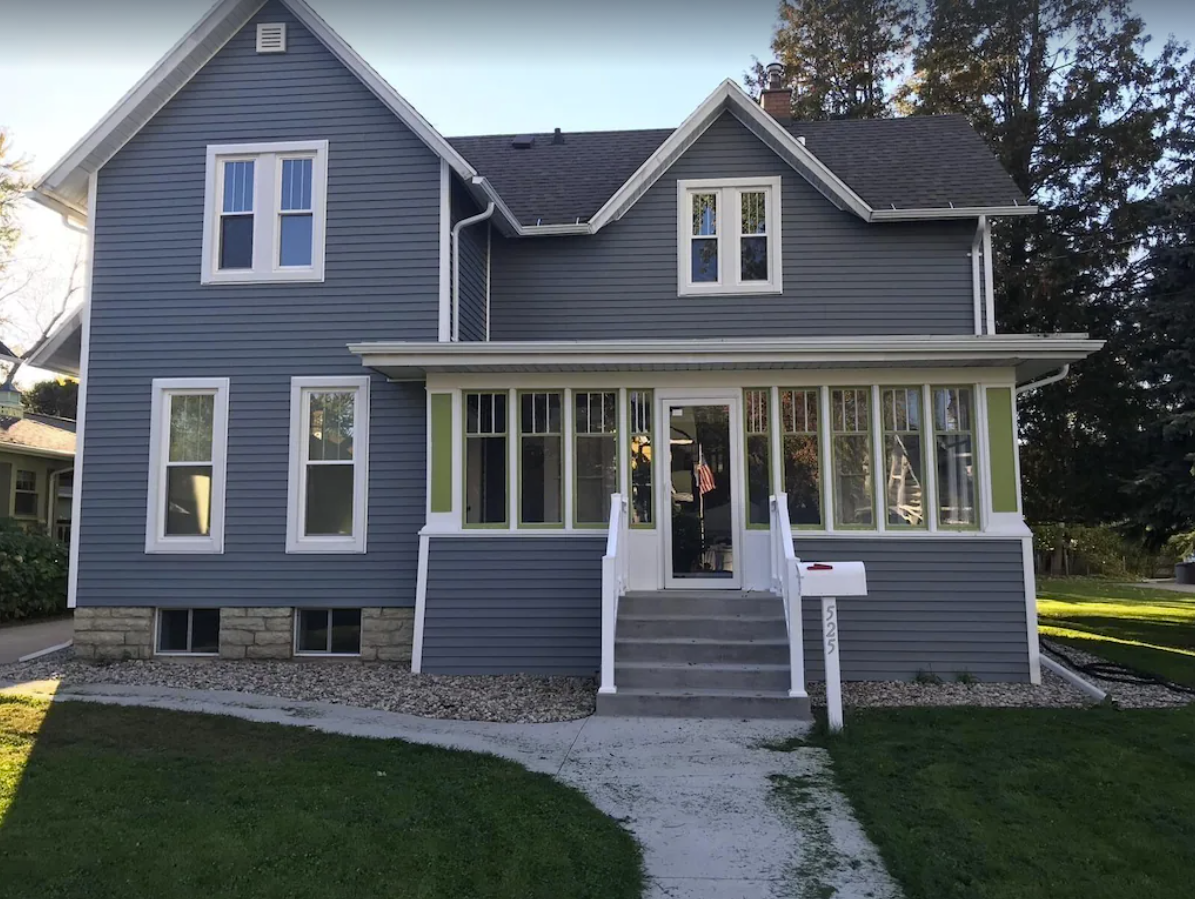 An exterior shot of a quaint two-story home with blue siding. The home has a small sunroom and white trimmed windows across the front of the home.