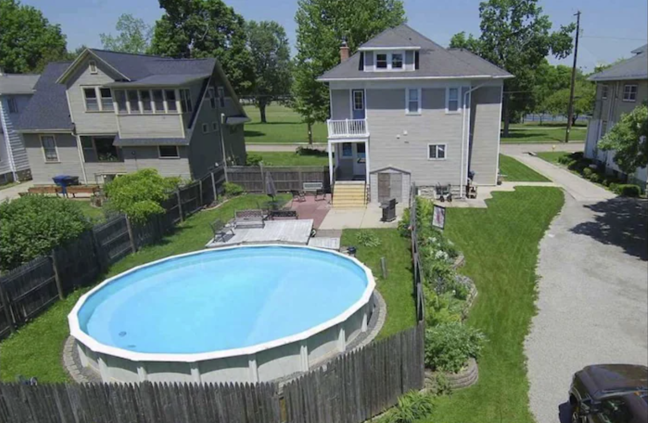 An exterior aerial shot of the backyard of a two-story home with an above-ground pool. The homes in the image both have the same gray-colored siding and lush yards with trees and shrubbery.