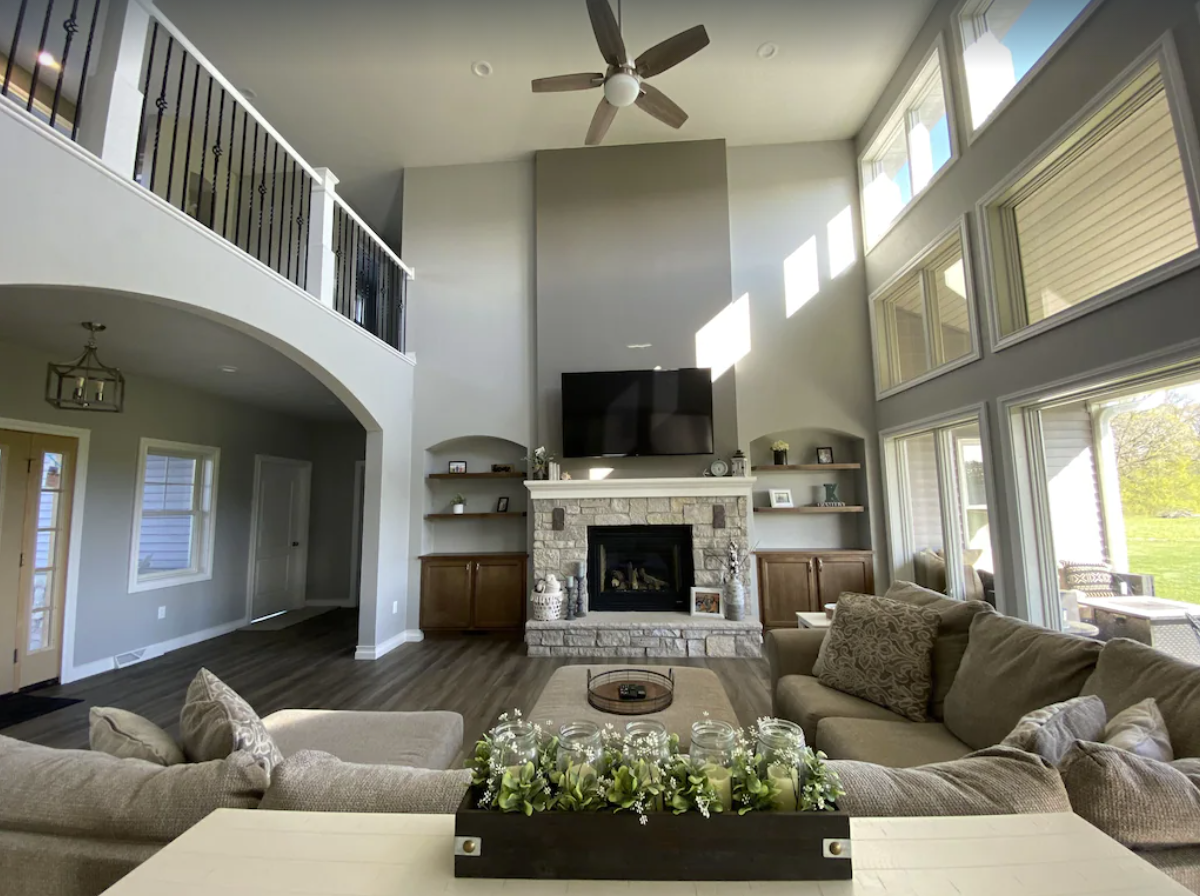 An elegant living room area with high ceilings, a large brown sectional couch, and stone fireplace. The floor-to-ceiling walls let in a lot of natural light, and a railing lines the open hallway on the second floor.