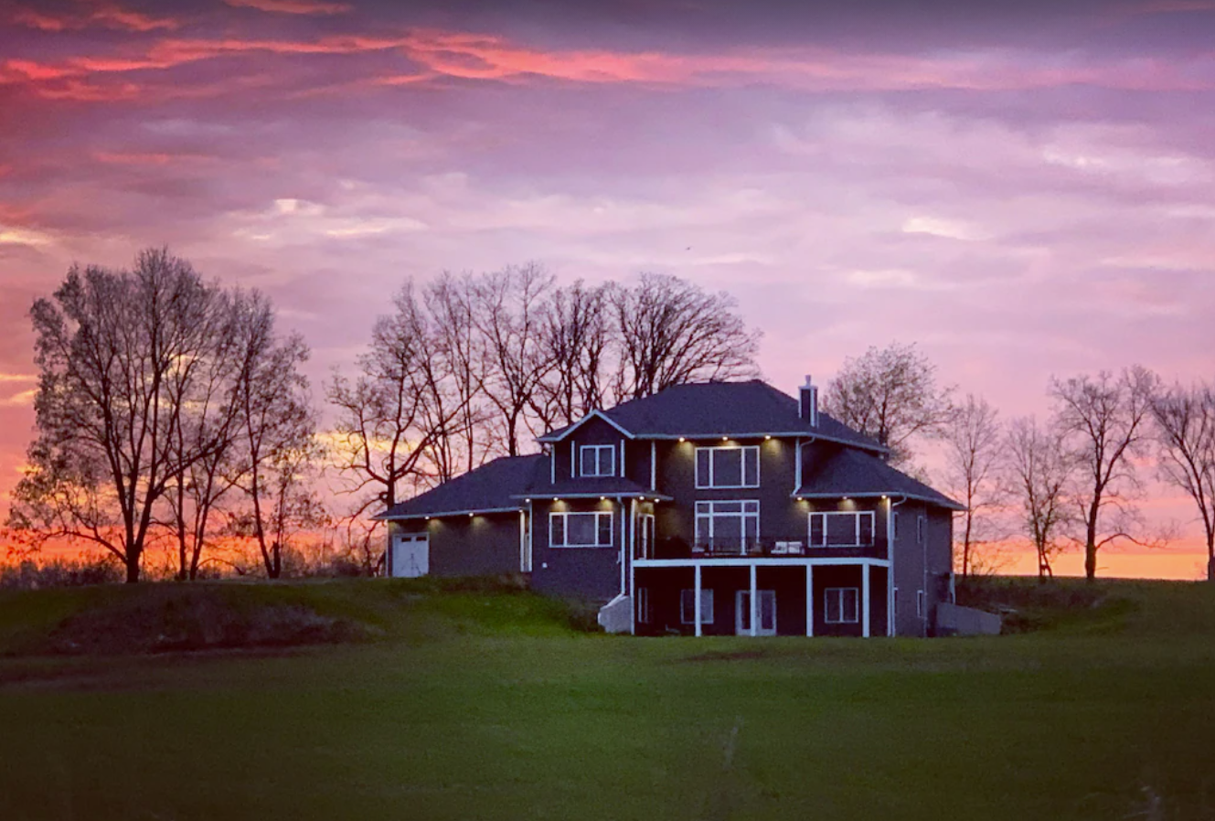 An exterior image shows a large three-story home against a pink and orange sunset sky. The home has dark gray siding and white trimmed windows across the back of the home, and an elevated balcony patio.