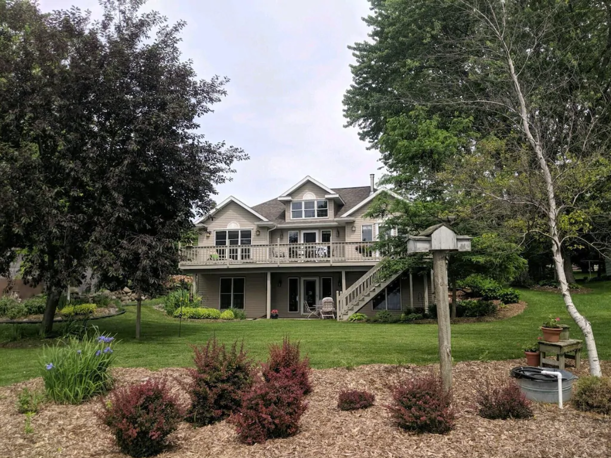 An exterior image of a large three story home and surrounding lawn. The home has a large wrap-around porch and staircase leading up to the second story. The home has tan and gray siding and white trim. The lawn is perfectly manicured with trees, mulch and bushes.