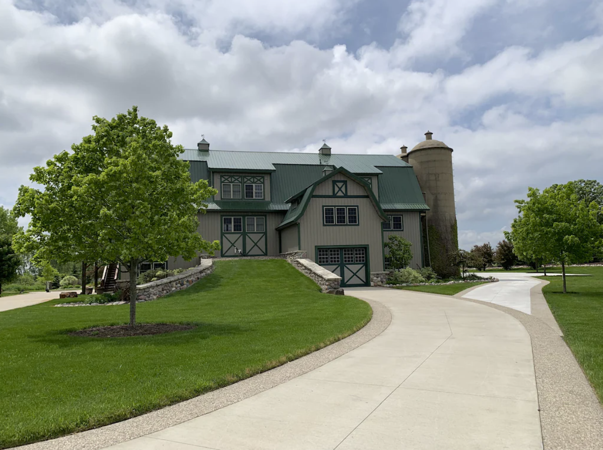 An exterior image of a large Oshkosh home. The house is a repurposed barn with gray and green exterior siding. A large concrete silo is attached to the side of the home. There's a long paved driveway leading up to a barndoor garage and manicured lawn.