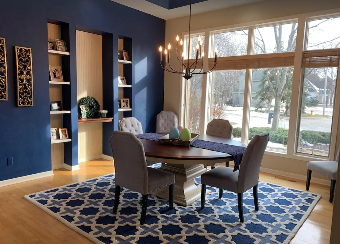 An interior shot of an elegant dining room with a navy blue accent wall and built-in display shelving. A round dining table has four seats, and sits on a blue area rug.