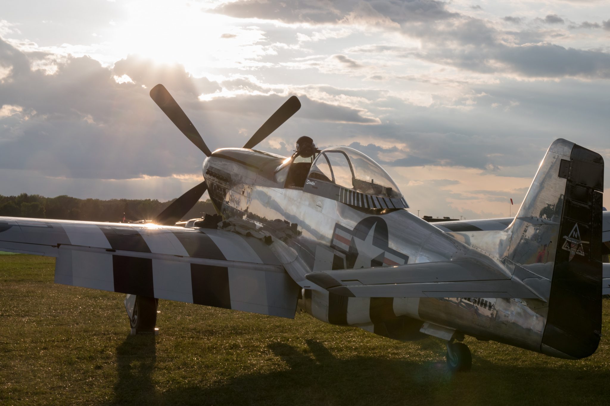 An old vintage plane sits in a grassy field at the EAA AirVenture air show. The sun shines through clouds in the sky across the horizon.