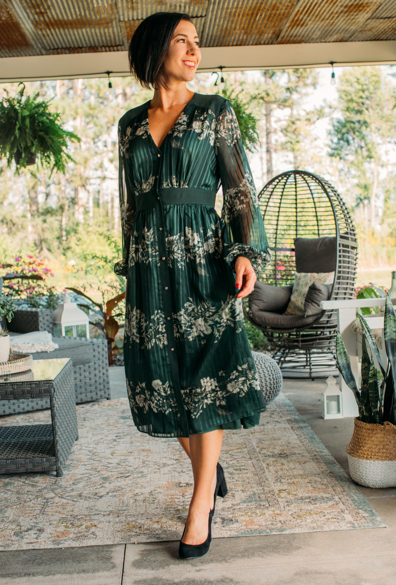 A woman poses in a long-sleeved green silk dress. The dress has a pleated pattern with white flowers, long and flowy translucent sleeves, and a v-neckline.