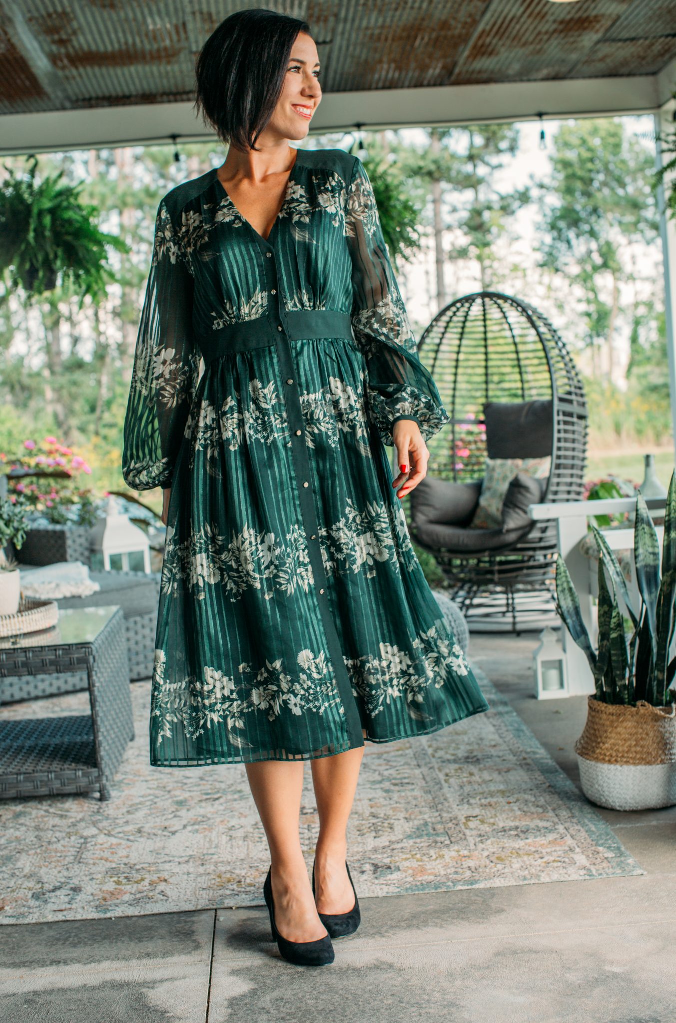A woman poses for the camera wearing a knee-length green silk dress with a white floral pattern. The dress has a pleated texture and has long, flowing sleeves.