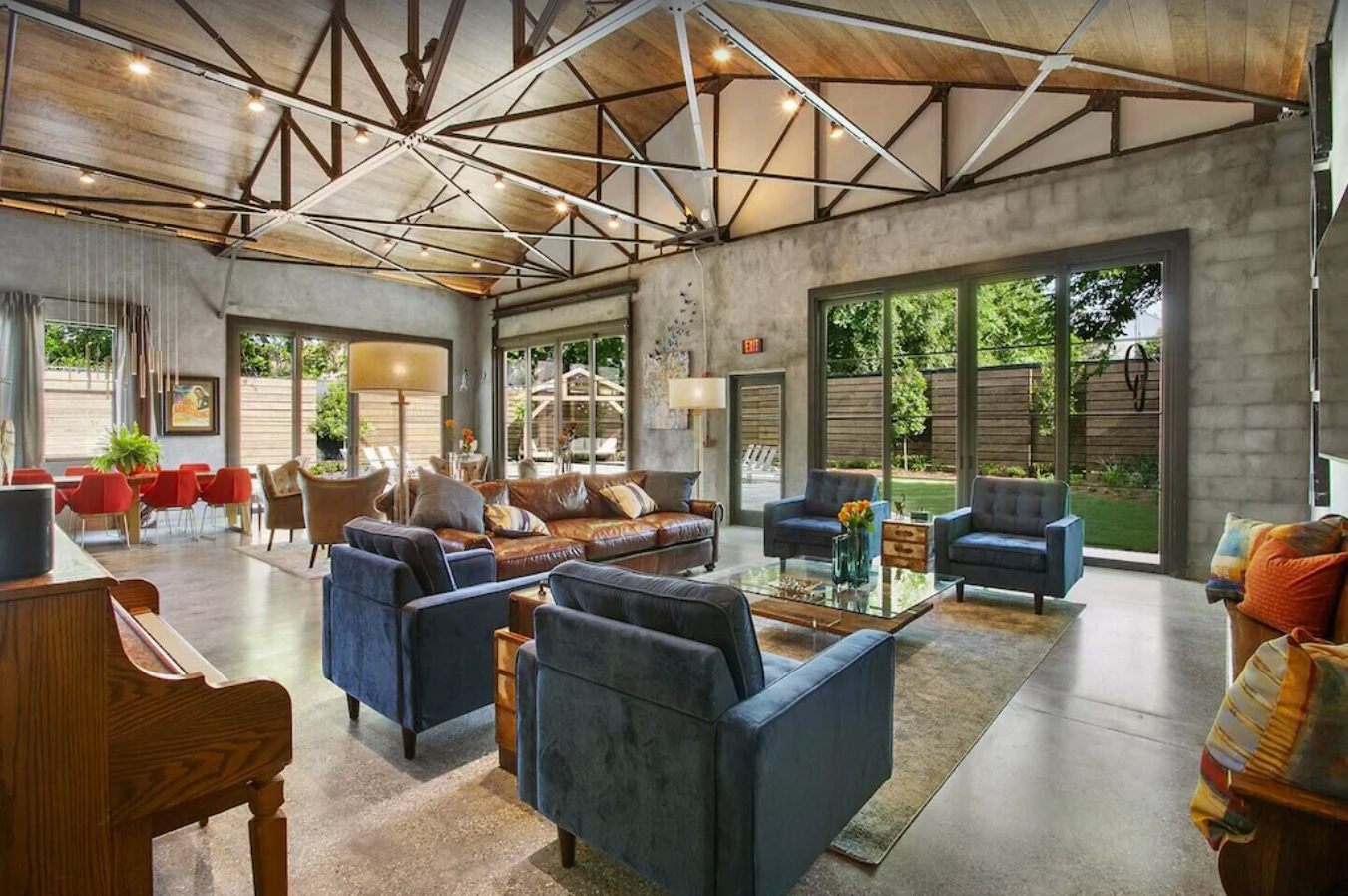 A modern living room decorated with mid-century style furniture. The walls are concrete gray with floor to ceiling windows, high wood paneled ceilings and beans, and concrete floors and area rugs around the furniture.