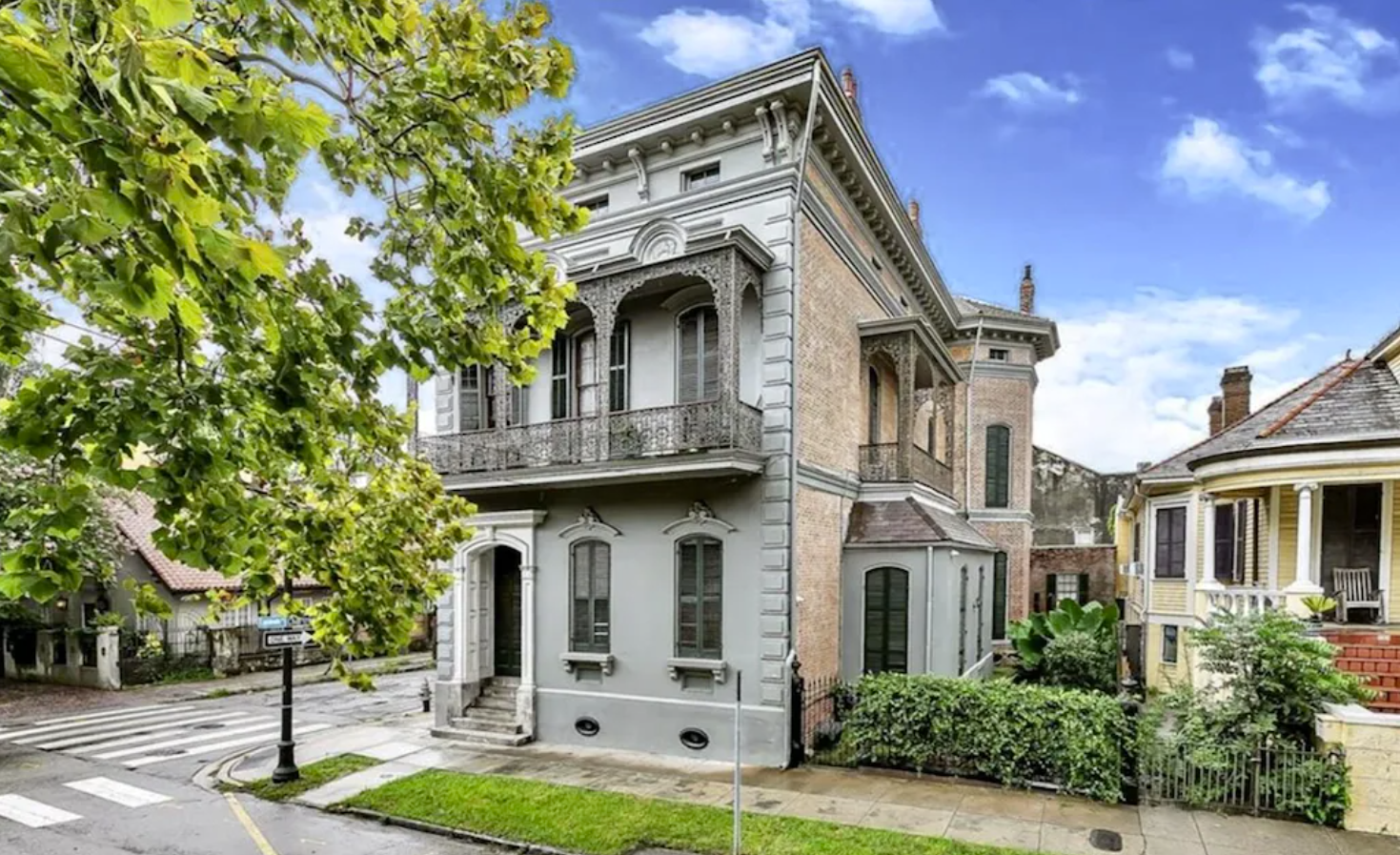 AN exterior image of a large two-story mansion on the corner in New Orleans. The front façade is gray concrete, and the side is a lighter, tan stone. Windows cover the front and sides of the building, and there's a small private patio on the side of the home.