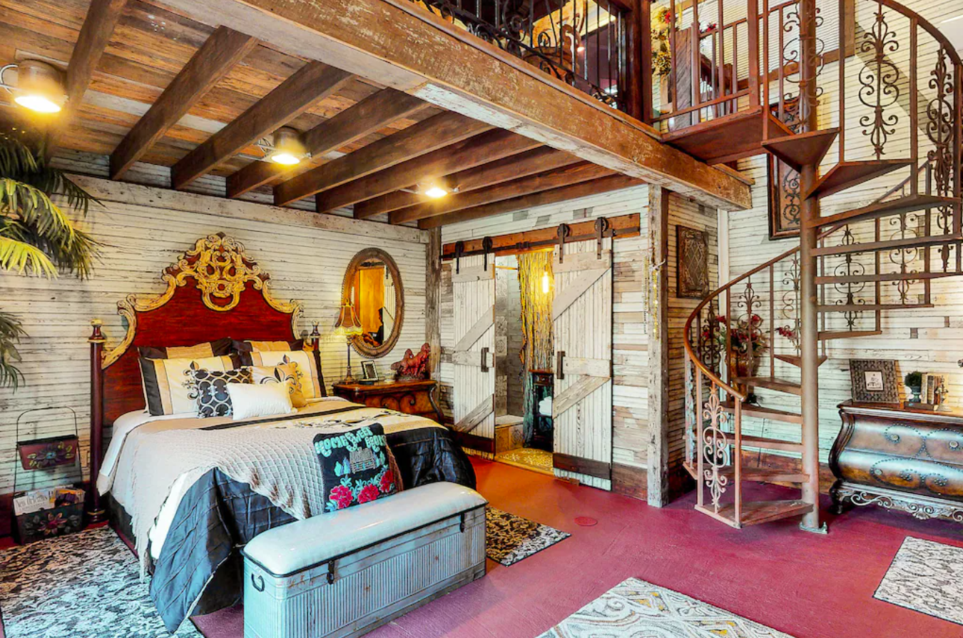 An interior image of a bedroom inside the Crab Shack loft in New Orleans. There's a king-size bed along a shiplap wall. A brass spiral staircase leads up to a loft-style living room.
