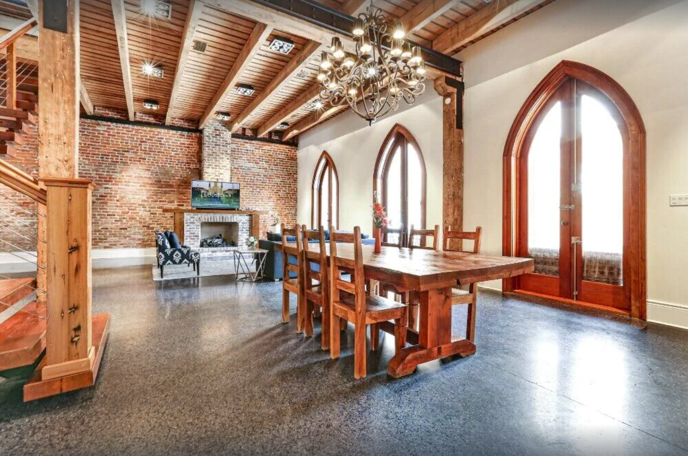 A gorgeous modern condo rental in New Orleans has an exposed brick wall and cathedral arched doorways. The ceiling is wood panels, and natural wood accents flow throughout the condo in a wooden dining table, staircase, and fireplace mantle.