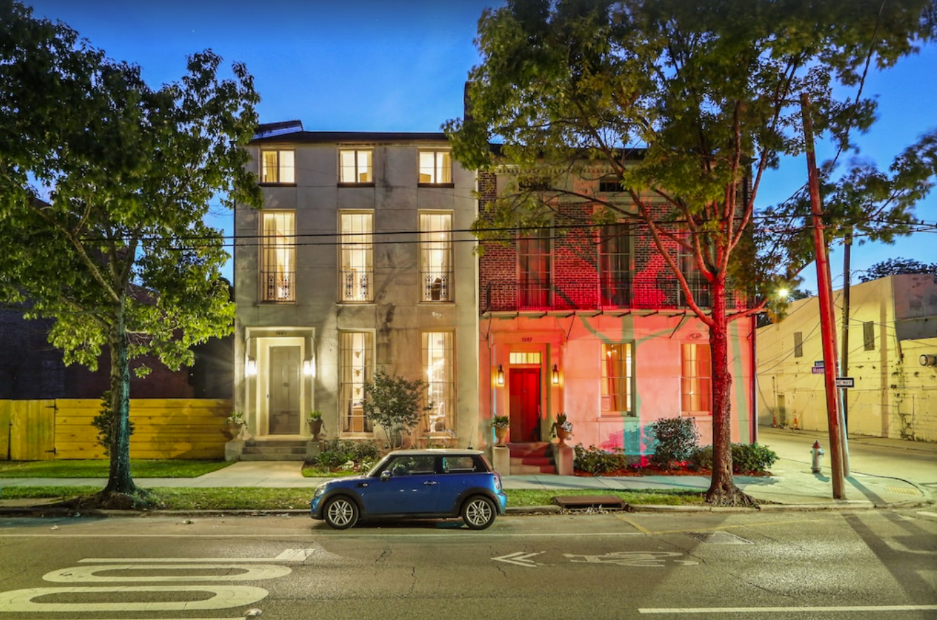 An exterior shot of two side-by-side homes in New Orleans. The front facades are both concrete, both two stories high. The home on the right is lit by red flood lights. A small blue fiat is parked out front.