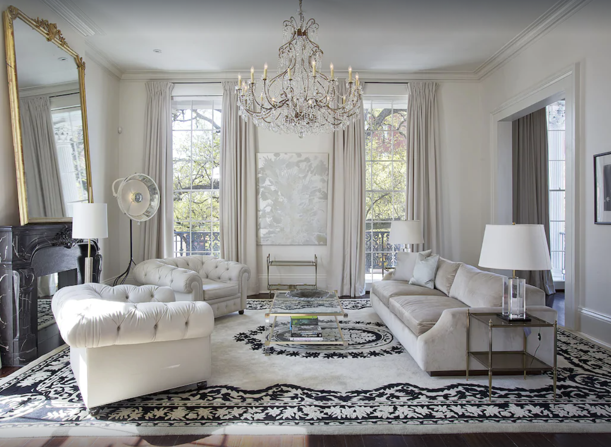 An interior shot of an elegant living room in a New Orleans home rental. The decor is luxurious and white, with white couches, a black and white area rug, white walls, curtains, and a grand chandelier. 