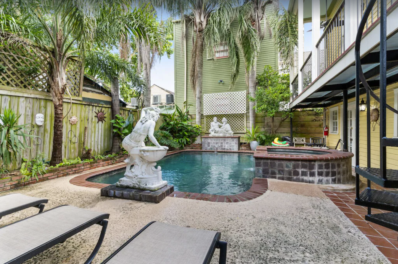 An image of an outdoor courtyard that's fenced in with a privacy fence. An in-ground pool and hot tub are the focal point of a concrete and brick patio. The house has a second story balcony with a spiral staircase.