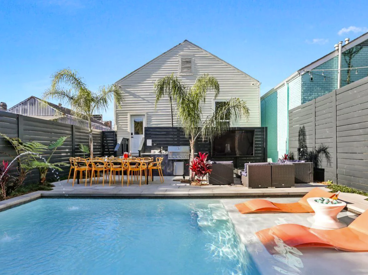 An image of an outdoor backyard oasis in New Orleans features a large in-ground pool with a deep end and shallow lounge area with chairs, a bar, patio furniture, and outdoor kitchen area.