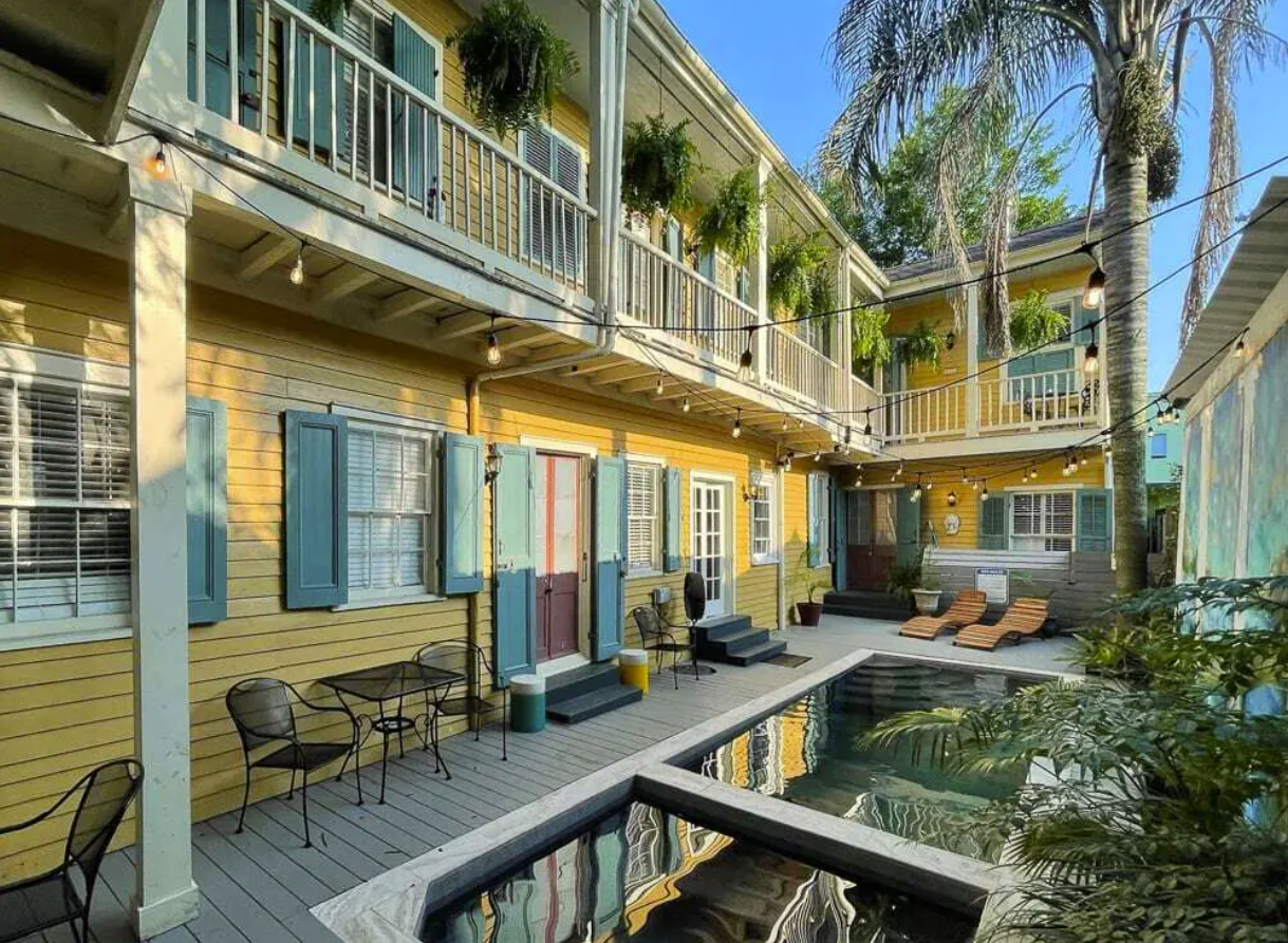 The backyard of a long, two-story VRBO rental in New Orleans. The home has a second floor balcony with hanging planters, yellow siding, and patio furniture on the deck. There's a side-by-side pool and hot tub on the deck as well.