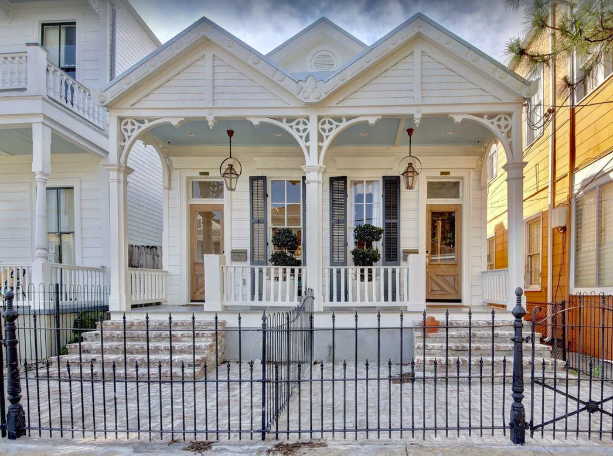 An exterior image of the outside of a New Orleans home. The side-by-side duplex has two entrances joined by a front porch. The white façade is single story and has a fenced-in stone-paved front yard.