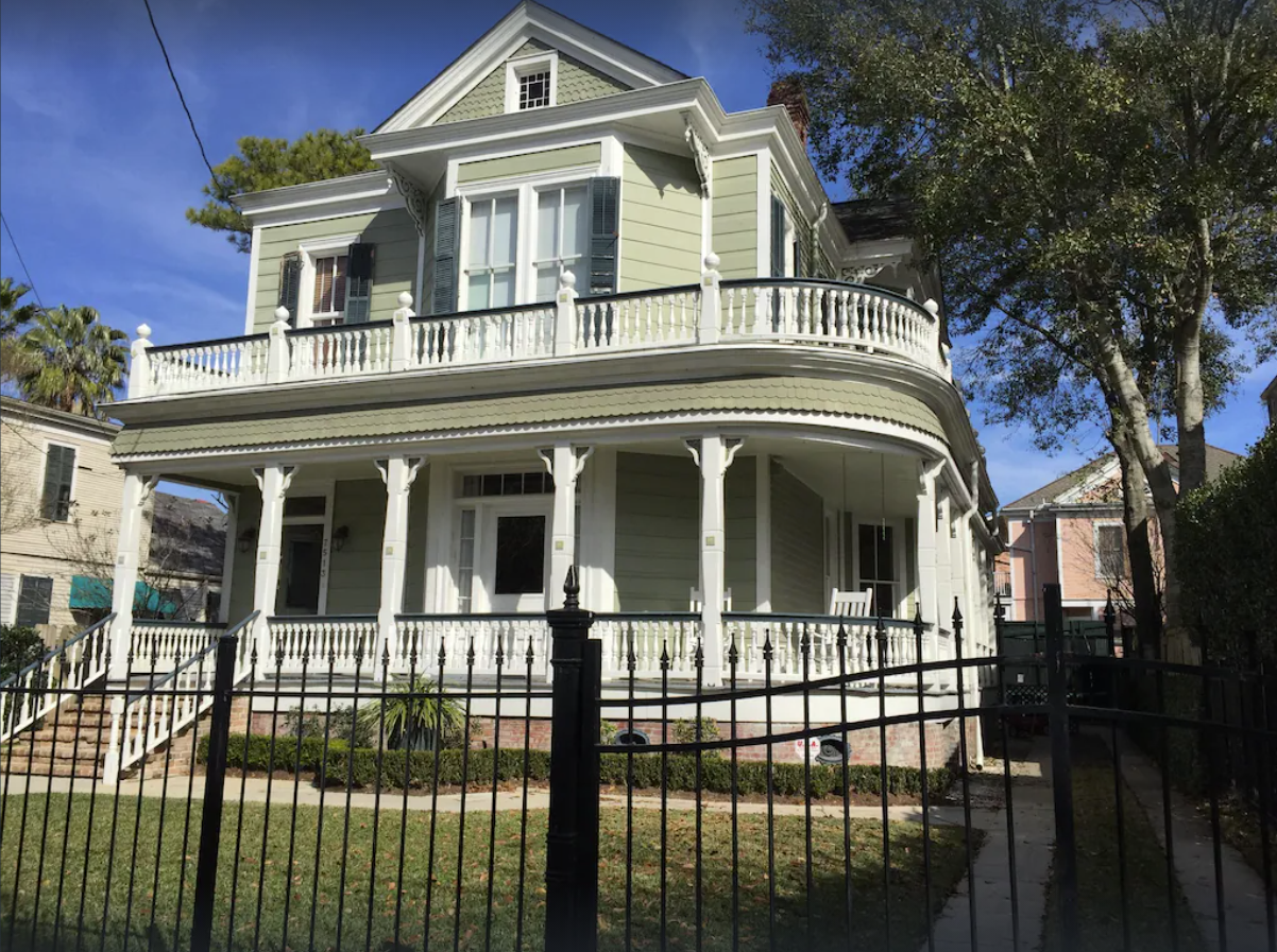An exterior image of the outside of a large two-story Queen Anne style historical home. The two-story home has a wrap-around front porch with light olive green siding and white pillars and a large fenced in yard.