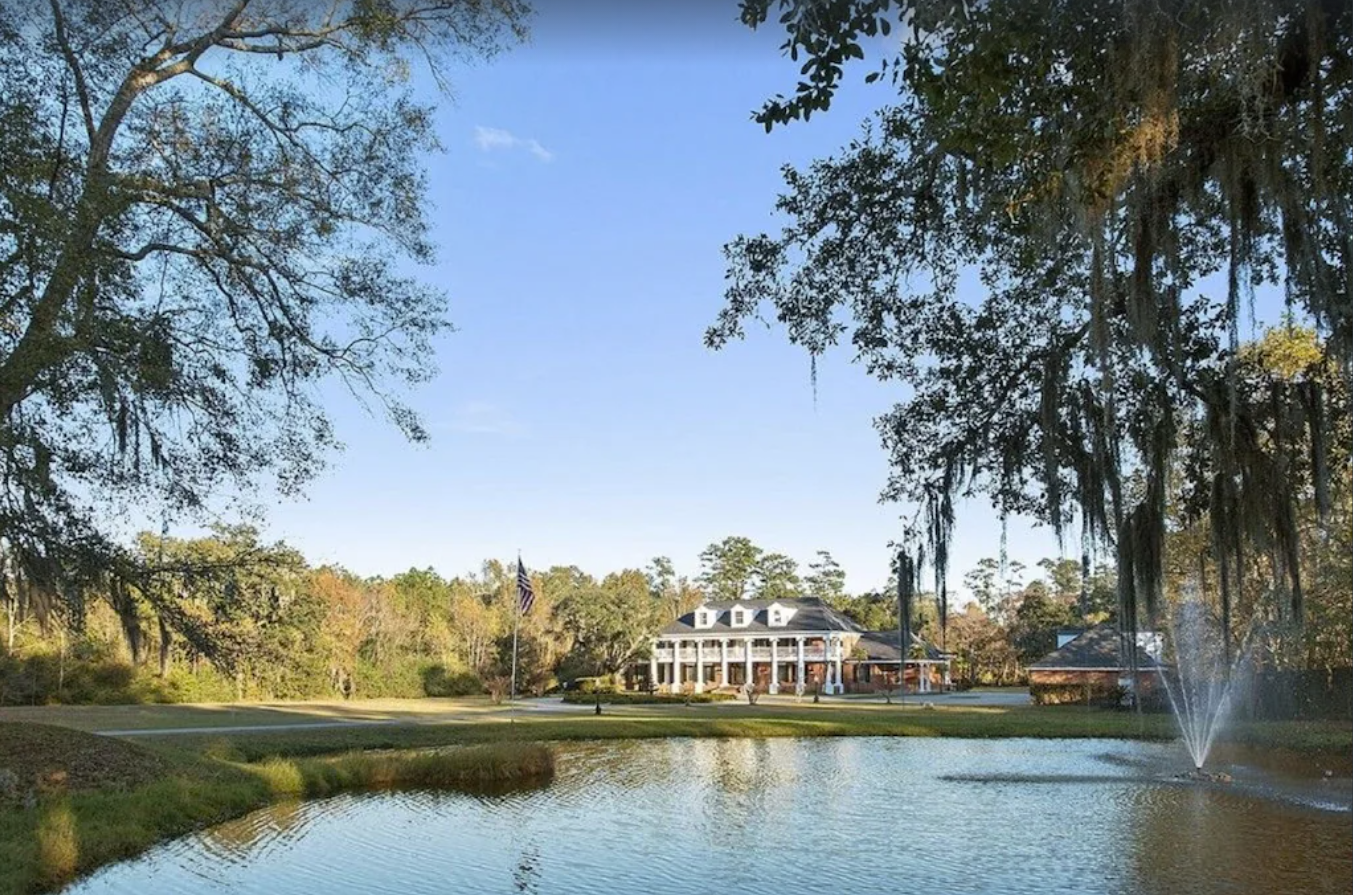 A gorgeous exterior image of a plantation property. There's a large pond surrounded by ancient oak trees, a manicured lawn, and in the background, a large two-story plantation home.