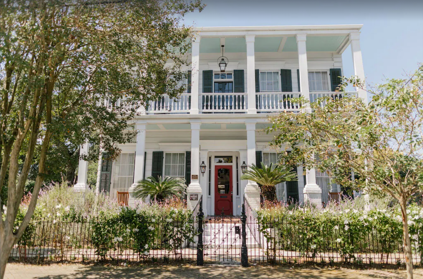 The outer front facade of a colonial style two-story home in New Orleans. The front yard has tall trees and vegetation. The home has a warp-around balcony on both stories, and a black wrought-iron gate around the property.