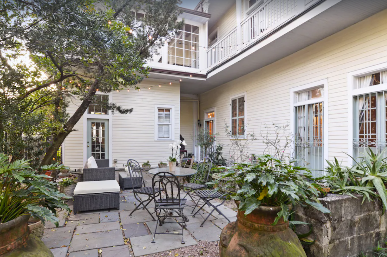 The back courtyard of a quaint two-story townhome in New Orleans. The siding and window trim are an eggshell white, and the stone patio is furnished with black patio furniture. String lights hang across the courtyard, strung from the balconies. 
